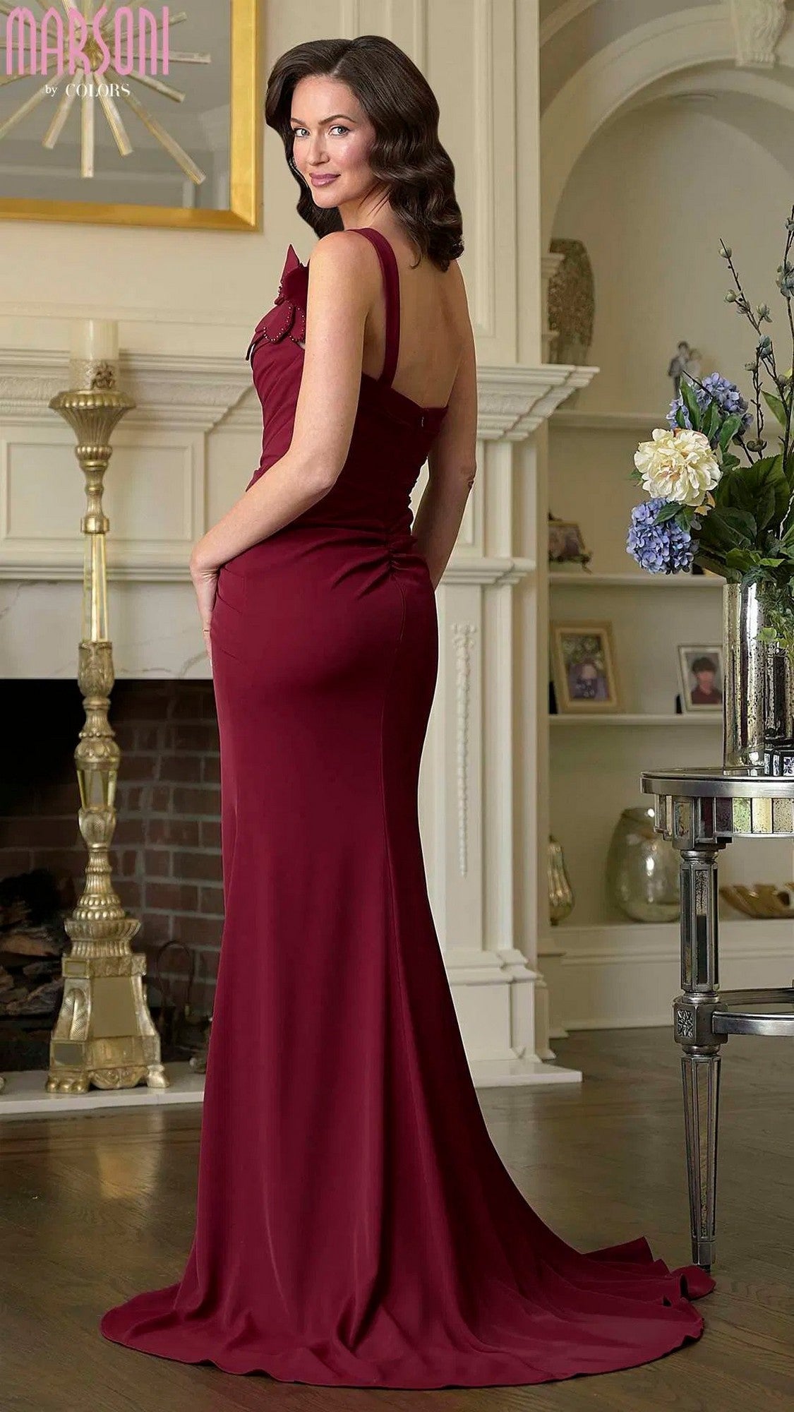 A woman wearing the Marsoni M330 Long Formal Dress poses in a stylish living room, standing with her hands in her pockets and glancing over her shoulder.